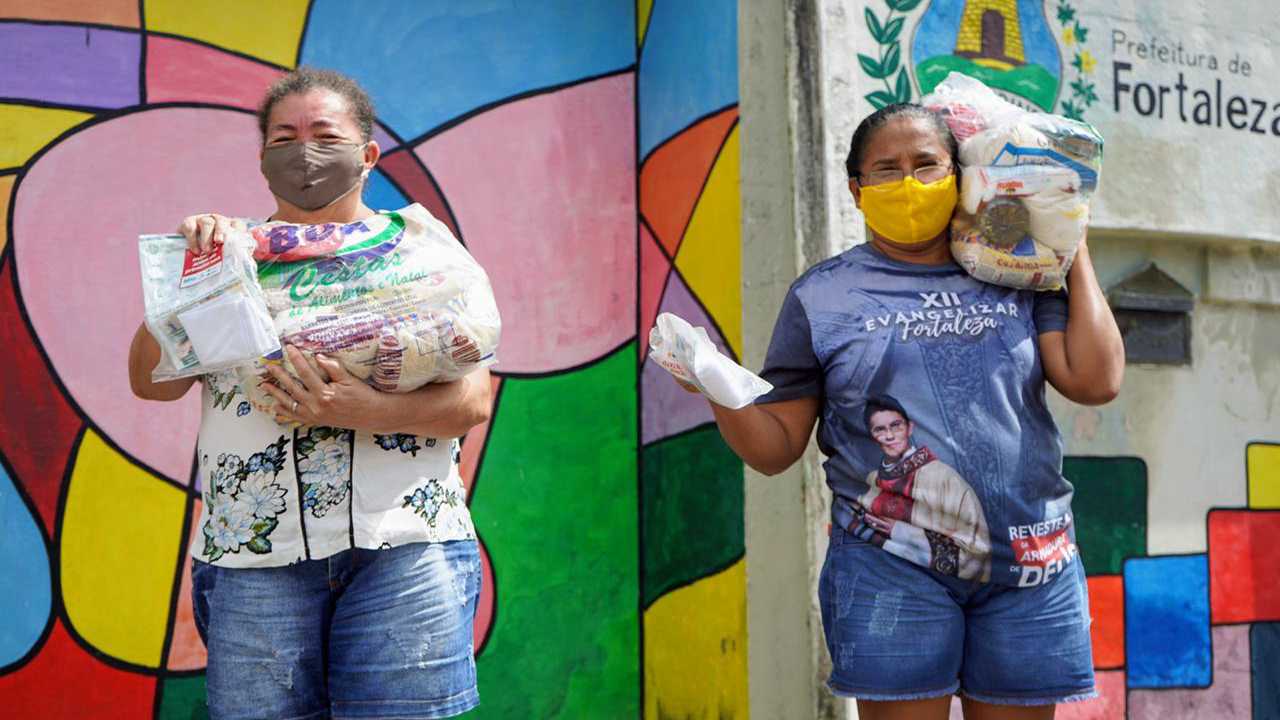 duas mulheres lado a lado posando para a foto de máscara e segurando cestas básicas